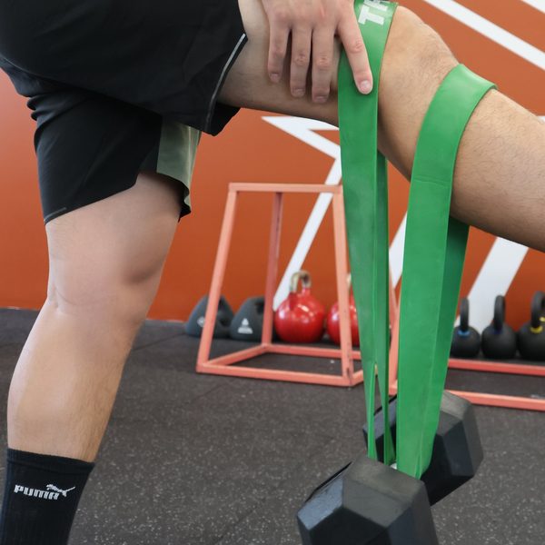 A green resistance band looped behind the knee and anchored to a dumbbell on the floor